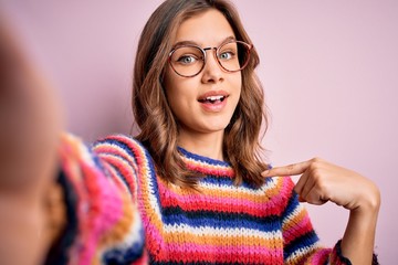 Young blonde girl wearing glasses taking a selfie picture of herself over pink isolated background with surprise face pointing finger to himself