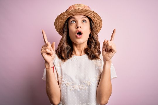 Young beautiful blonde girl wearing summer hat over pink isolated background amazed and surprised looking up and pointing with fingers and raised arms.