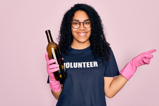 African american curly woman wearing volunteer t-shirt doing volunteering recycling glass bottle very happy pointing with hand and finger to the side