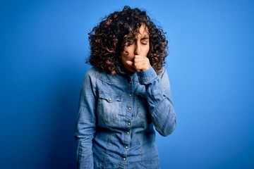 Young beautiful curly arab woman wearing casual denim shirt standing over blue background feeling unwell and coughing as symptom for cold or bronchitis. Health care concept.