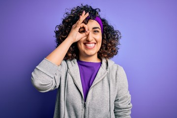 Beautiful curly arab sportswoman doing sport wearing sportswear over purple background doing ok gesture with hand smiling, eye looking through fingers with happy face.