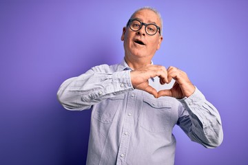 Middle age handsome hoary man wearing striped shirt and glasses over purple background smiling in love doing heart symbol shape with hands. Romantic concept.