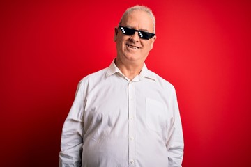 Middle age hoary man wearing funny sunglasses over isolated red background with a happy and cool smile on face. Lucky person.