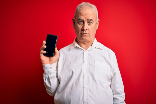 Middle age hoary man holding smartphone showing screen over isolated red background with a confident expression on smart face thinking serious