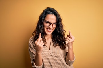 Beautiful woman with curly hair wearing striped shirt and glasses over yellow background Showing middle finger doing fuck you bad expression, provocation and rude attitude. Screaming excited