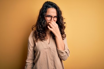Beautiful woman with curly hair wearing striped shirt and glasses over yellow background smelling something stinky and disgusting, intolerable smell, holding breath with fingers on nose. Bad smell
