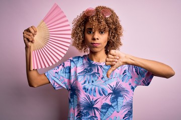 Young african american curly woman on vacation using hand fan over isolated pink background with angry face, negative sign showing dislike with thumbs down, rejection concept