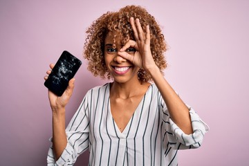 Young african american curly woman holding broken smartphone showing craked screen with happy face...