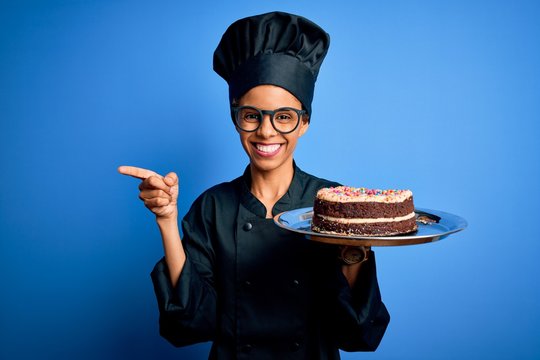 Young african american baker woman wearing cooker uniform and hat holding cake very happy pointing with hand and finger to the side