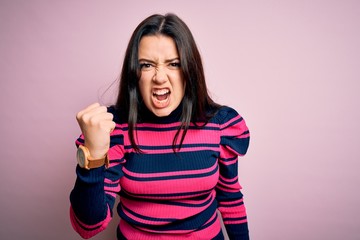 Young brunette elegant woman wearing striped shirt over pink isolated background angry and mad raising fist frustrated and furious while shouting with anger. Rage and aggressive concept.