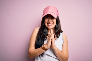 Young brunette woman wearing casual sport cap over pink background praying with hands together asking for forgiveness smiling confident.