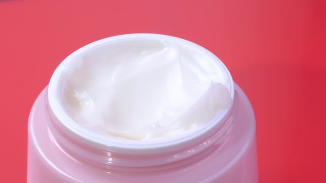 A woman takes a white hand cream from a pink jar with her finger. The concept of beauty and health and skin care for the body, face and legs. Macro. Close up view on a red background