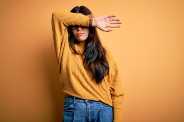 Young brunette woman wearing glasses and casual sweater over yellow isolated background covering eyes with arm, looking serious and sad. Sightless, hiding and rejection concept