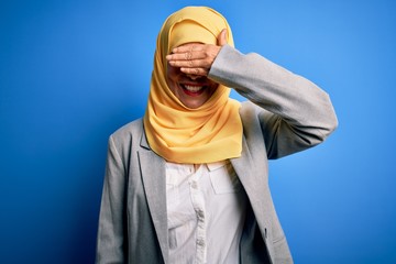 Fototapeta na wymiar Middle age brunette business woman wearing muslim traditional hijab over blue background smiling and laughing with hand on face covering eyes for surprise. Blind concept.