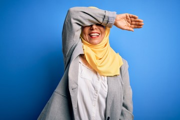 Fototapeta na wymiar Middle age brunette business woman wearing muslim traditional hijab over blue background covering eyes with arm smiling cheerful and funny. Blind concept.