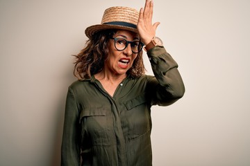 Middle age brunette woman wearing glasses and hat standing over isolated white background surprised...