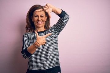 Middle age beautiful woman wearing casual sweater standing over isolated pink background smiling making frame with hands and fingers with happy face. Creativity and photography concept.