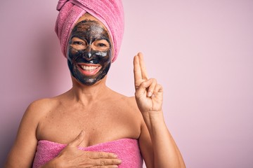 Middle age brunette woman wearing beauty black face mask over isolated pink background smiling swearing with hand on chest and fingers up, making a loyalty promise oath