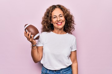 Middle age beautiful sporty woman playing rugby holding football ball over pink background looking positive and happy standing and smiling with a confident smile showing teeth - Powered by Adobe
