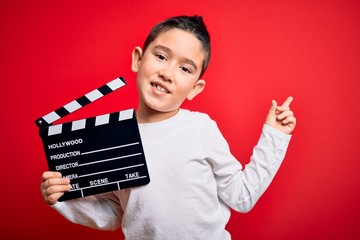 Young little boy kid filming video holding cinema director clapboard over isolated red background...