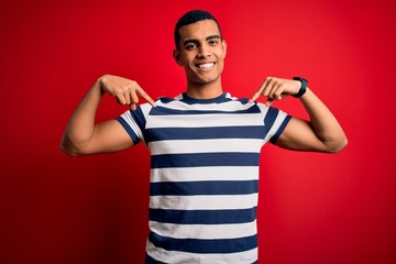 Handsome african american man wearing casual striped t-shirt standing over red background looking confident with smile on face, pointing oneself with fingers proud and happy.