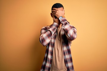 Young handsome african american man wearing casual shirt standing over yellow background Covering eyes and mouth with hands, surprised and shocked. Hiding emotion