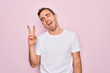 Handsome man with blue eyes wearing casual white t-shirt standing over pink background smiling with happy face winking at the camera doing victory sign. Number two.