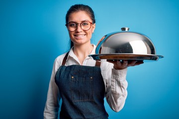 Young beautiful waitress woman with blue eyes holding tray with dome over isolated background with a happy face standing and smiling with a confident smile showing teeth
