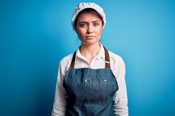 Young beautiful baker woman with blue eyes wearing apron and cap over blue background with serious expression on face. Simple and natural looking at the camera.