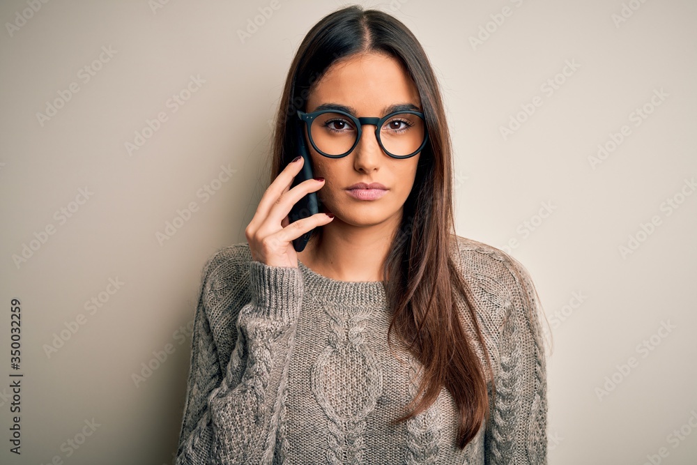 Wall mural Young beautiful brunette woman wearing glasses having conversation talking on smartphone with a confident expression on smart face thinking serious