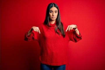 Young beautiful brunette woman wearing casual sweater over isolated red background Pointing down looking sad and upset, indicating direction with fingers, unhappy and depressed.