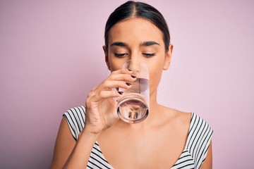 Young beautiful brunette woman drinking glass of healthy water to refreshment standing over isolated pink background