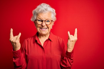Senior beautiful grey-haired woman wearing casual shirt and glasses over red background shouting with crazy expression doing rock symbol with hands up. Music star. Heavy concept.