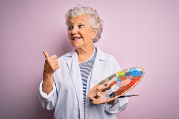 Senior beautiful grey-haired artist woman painting using brush and palette over pink background...