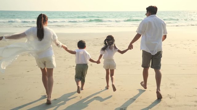 Slow motion of young Asian family parent and two child kid holding hands together and running on the beach with smiling at summer sunset. Happy family relax and having fun in holiday vacation travel.