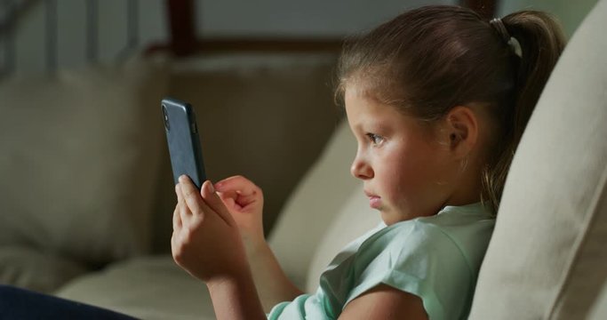 Authentic shot of a happy little girl is having fun to use a smartphone for playing games or study while sitting on a sofa in a living room at home.