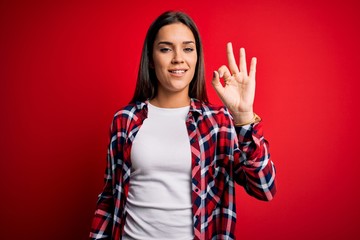 Young beautiful brunette woman wearing casual shirt standing over isolated red background smiling positive doing ok sign with hand and fingers. Successful expression.