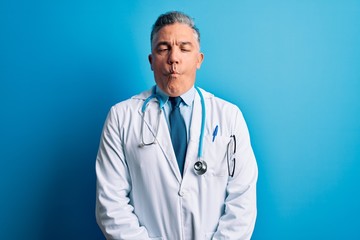 Middle age handsome grey-haired doctor man wearing coat and blue stethoscope making fish face with lips, crazy and comical gesture. Funny expression.
