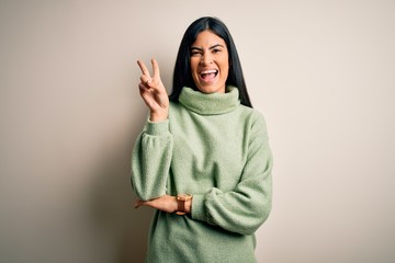 Young beautiful hispanic woman wearing green winter sweater over isolated background smiling with happy face winking at the camera doing victory sign with fingers. Number two.