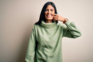 Young beautiful hispanic woman wearing green winter sweater over isolated background Pointing with hand finger to face and nose, smiling cheerful. Beauty concept