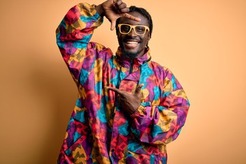 Handsome african american man wearing colorful coat and glasses over yellow background smiling making frame with hands and fingers with happy face. Creativity and photography concept.