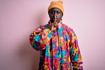 Young handsome african american man wearing colorful coat and cap over pink background asking to be quiet with finger on lips. Silence and secret concept.