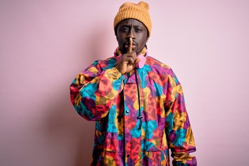 Obraz premium Young handsome african american man wearing colorful coat and cap over pink background asking to be quiet with finger on lips. Silence and secret concept.