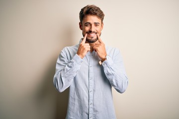 Young handsome man with beard wearing striped shirt standing over white background Smiling with open mouth, fingers pointing and forcing cheerful smile