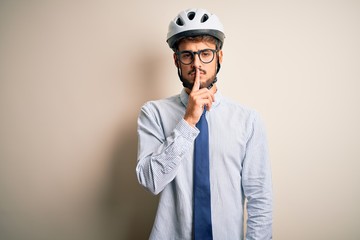 Young businessman wearing glasses and bike helmet standing over isolated white bakground asking to be quiet with finger on lips. Silence and secret concept.