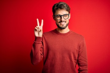 Young handsome man with beard wearing glasses and sweater standing over red background showing and pointing up with fingers number two while smiling confident and happy.