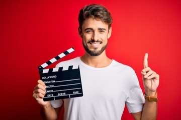 Young director man with beard making movie using clapboard over isolated red background surprised...