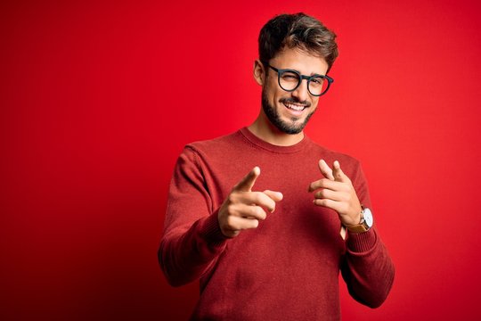 Young handsome man with beard wearing glasses and sweater standing over red background pointing fingers to camera with happy and funny face. Good energy and vibes.