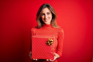 Young beautiful brunette woman holding birthday gift over isolated red background with a happy face standing and smiling with a confident smile showing teeth