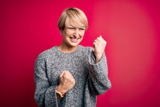 Young blonde woman with modern short hair wearing casual sweater over pink background celebrating surprised and amazed for success with arms raised and eyes closed. Winner concept.